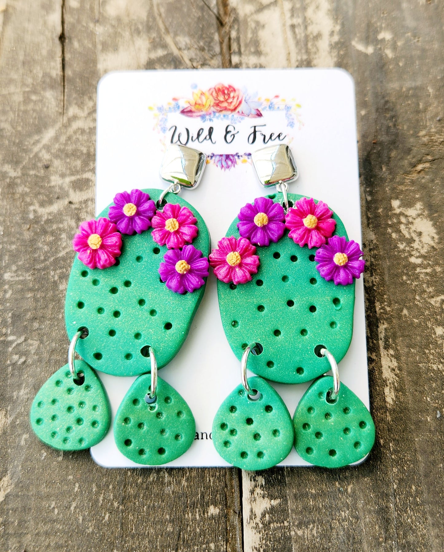 Handmade polymer clay desert cactus earrings! Lightweight! Unique design! A green cactus hangs from a silver-plated shiny square stud followed by two small cactus dangles. This cactus earring has four decorative flowers, two pink and two purple. The design has holes throughout to simulate cactus spines.