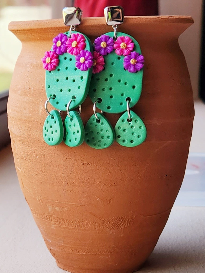 Handmade polymer clay desert cactus earrings! Lightweight! Unique design! A green cactus hangs from a silver-plated shiny square stud followed by two small cactus dangles. This cactus earring has four decorative flowers, two pink and two purple. The design has holes throughout to simulate cactus spines.