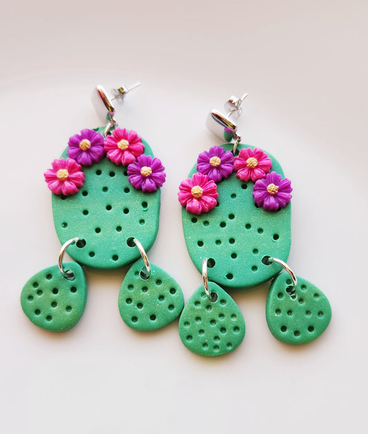 Handmade polymer clay desert cactus earrings! Lightweight! Unique design! A green cactus hangs from a silver-plated shiny square stud followed by two small cactus dangles. This cactus earring has four decorative flowers, two pink and two purple. The design has holes throughout to simulate cactus spines.