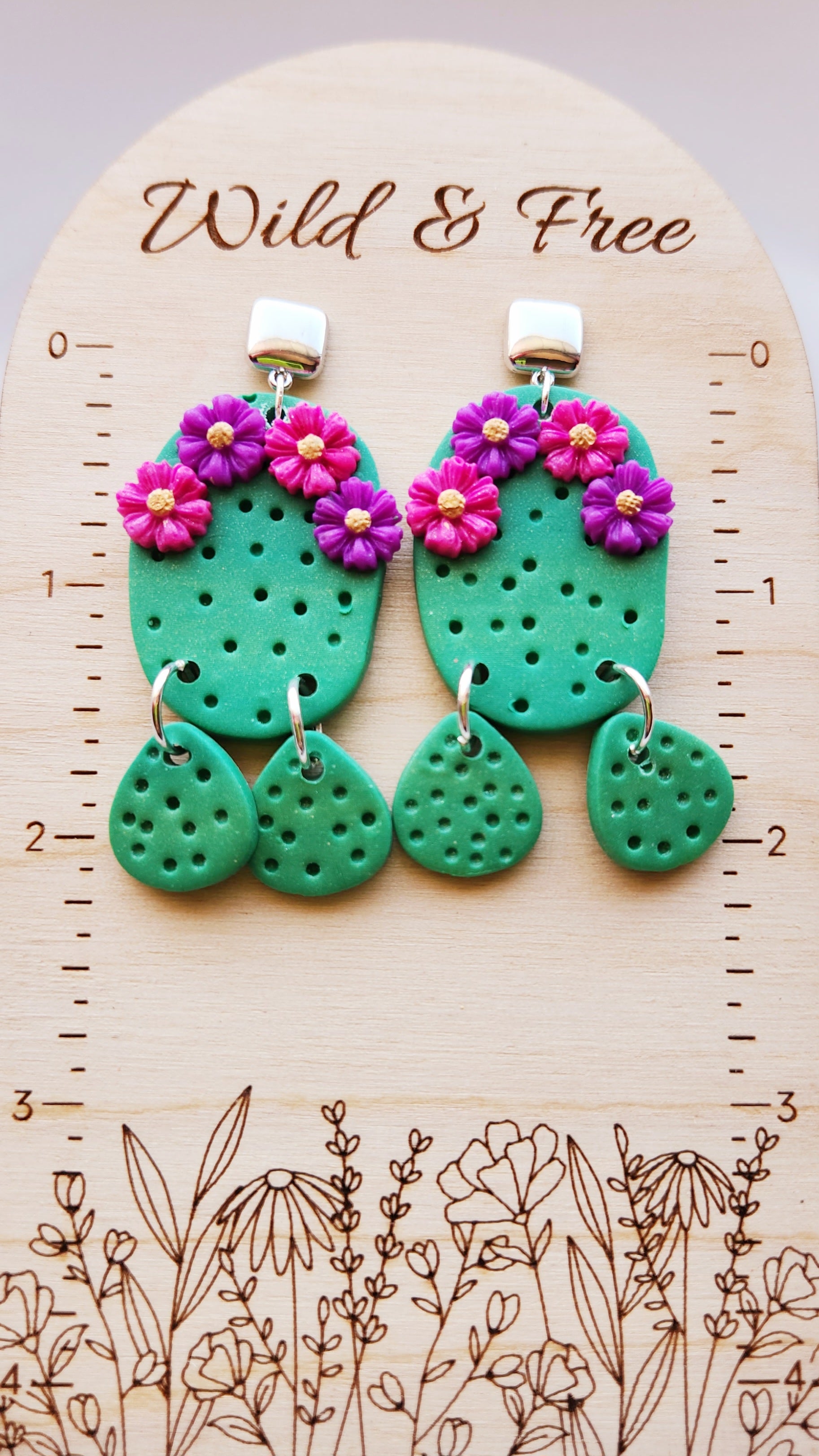 Handmade polymer clay desert cactus earrings! Lightweight! Unique design! A green cactus hangs from a silver-plated shiny square stud followed by two small cactus dangles. This cactus earring has four decorative flowers, two pink and two purple. The design has holes throughout to simulate cactus spines.