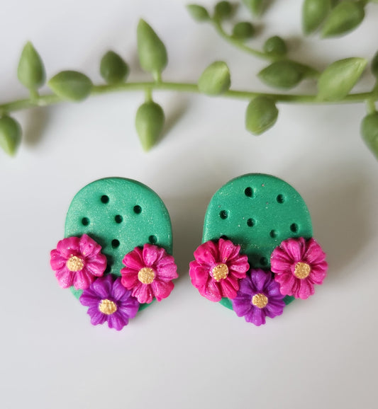 Handmade polymer clay desert cactus earrings! Lightweight! Unique design! A green cactus oval shape makes this stud design. This cactus earring has three decorative flowers, two pink and one purple. The design has holes throughout to simulate cactus spines. Lightweight, matte and approximately 1" long.