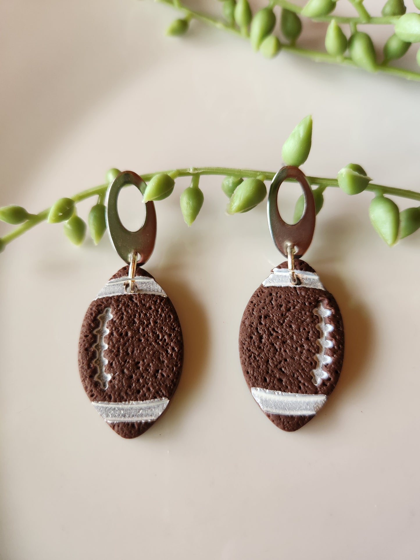 Handmade polymer clay football earrings! Be gameday-ready with these beautiful football earrings! No matter what your team is you are sure to stand out with these lightweight football dangles. These earrings are cut like a football shape with brown color clay. White paint highlights the ball design. They are textured matte with silver-plated open oval studs. Approximately 2" long.